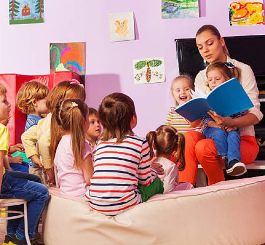 Large group of kids sit and listen to teacher reading a book and telling stories